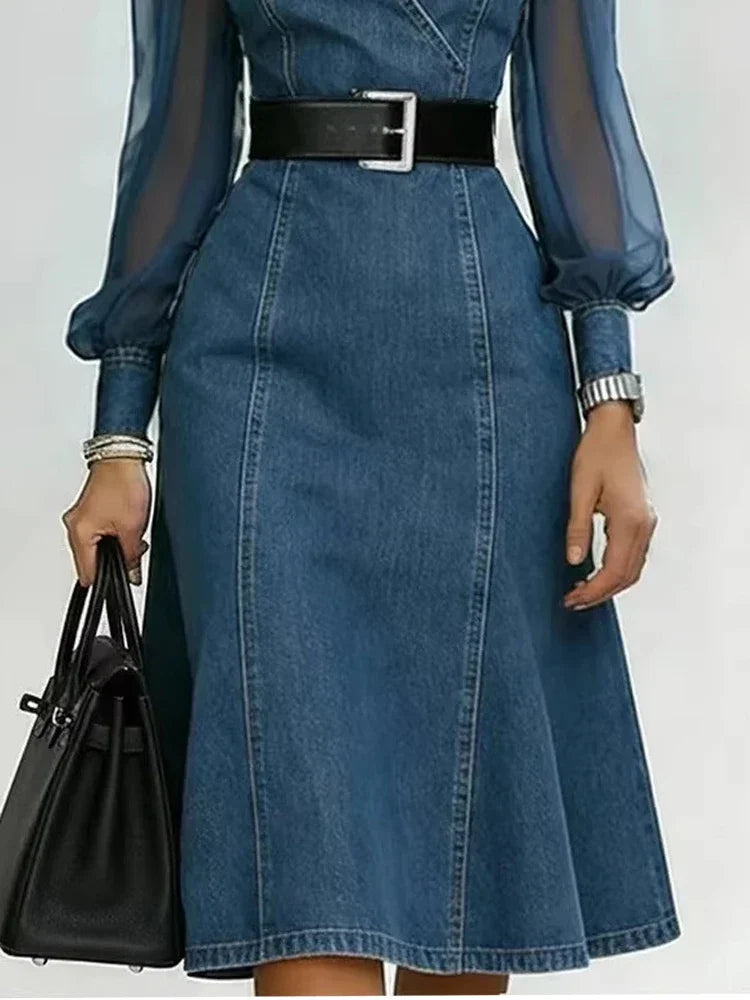 A woman wearing a blue denim midi dress with sheer long sleeves, a black belt with a silver buckle, holding a black handbag against a light gray background.