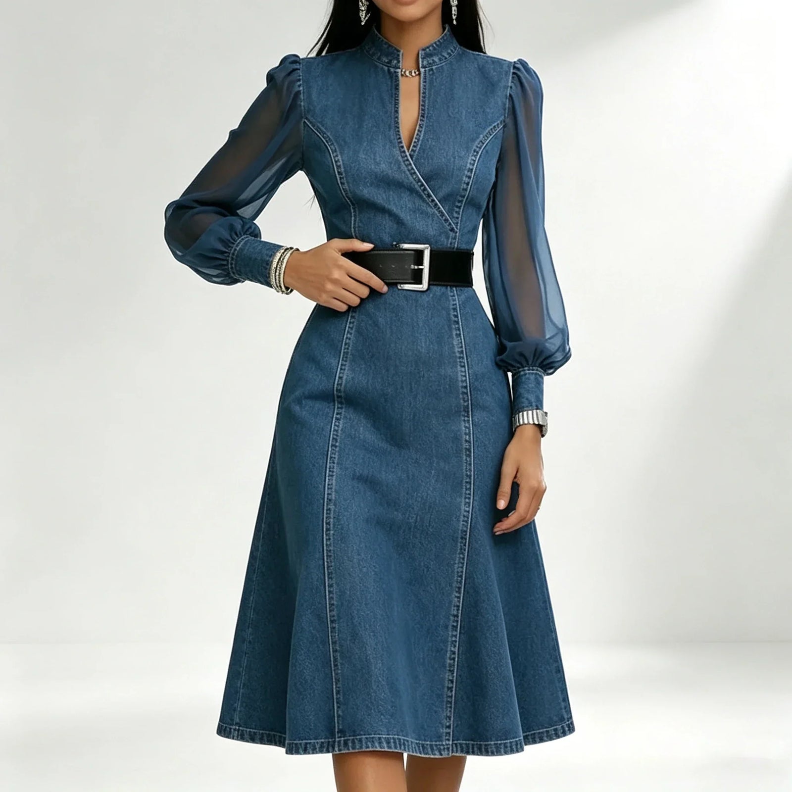 A woman wearing a blue denim midi dress with sheer long sleeves, a split neckline, and a black belt with a silver buckle, photographed against a light gray studio background.