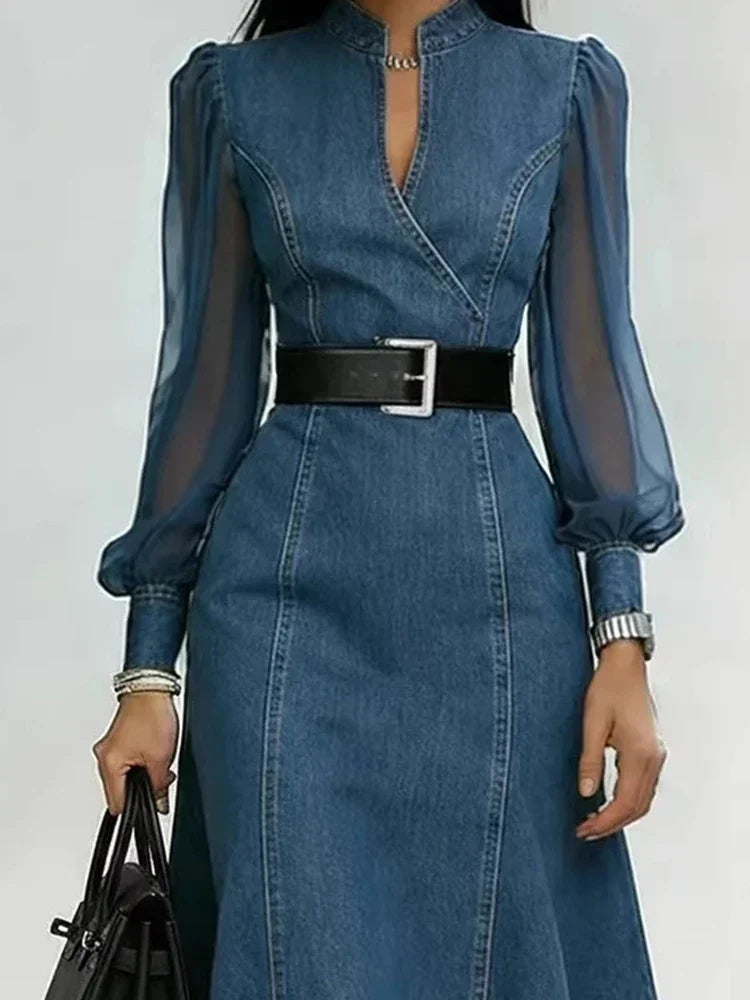 A woman wearing a blue denim midi dress with sheer long sleeves, a split neckline, and a black belt with a silver buckle, holding a black handbag against a plain light gray background.