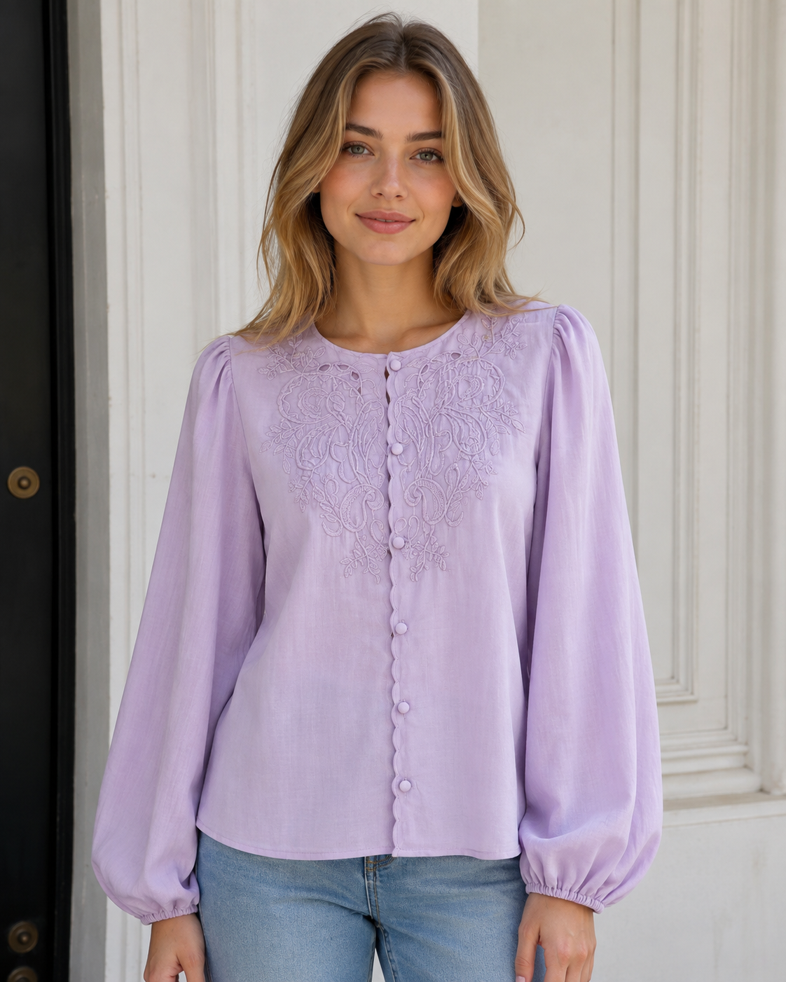 A woman wearing a lavender long-sleeve blouse with floral embroidery and a button-up front, paired with blue jeans, standing in front of a white paneled door.