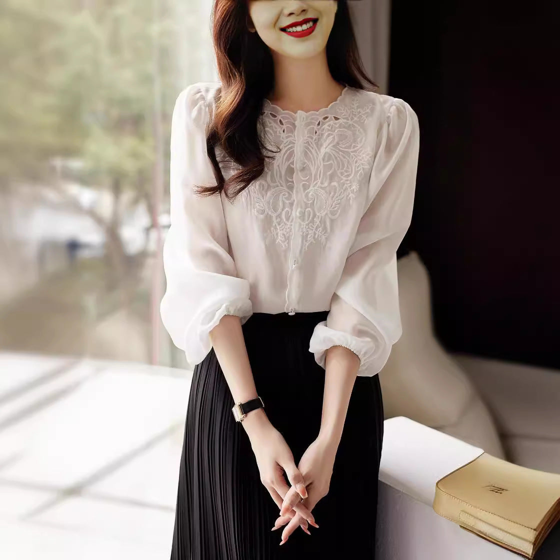 A woman wearing a white long-sleeve blouse with a sheer lace embroidered yoke and black pleated skirt, standing in a softly lit indoor setting.
