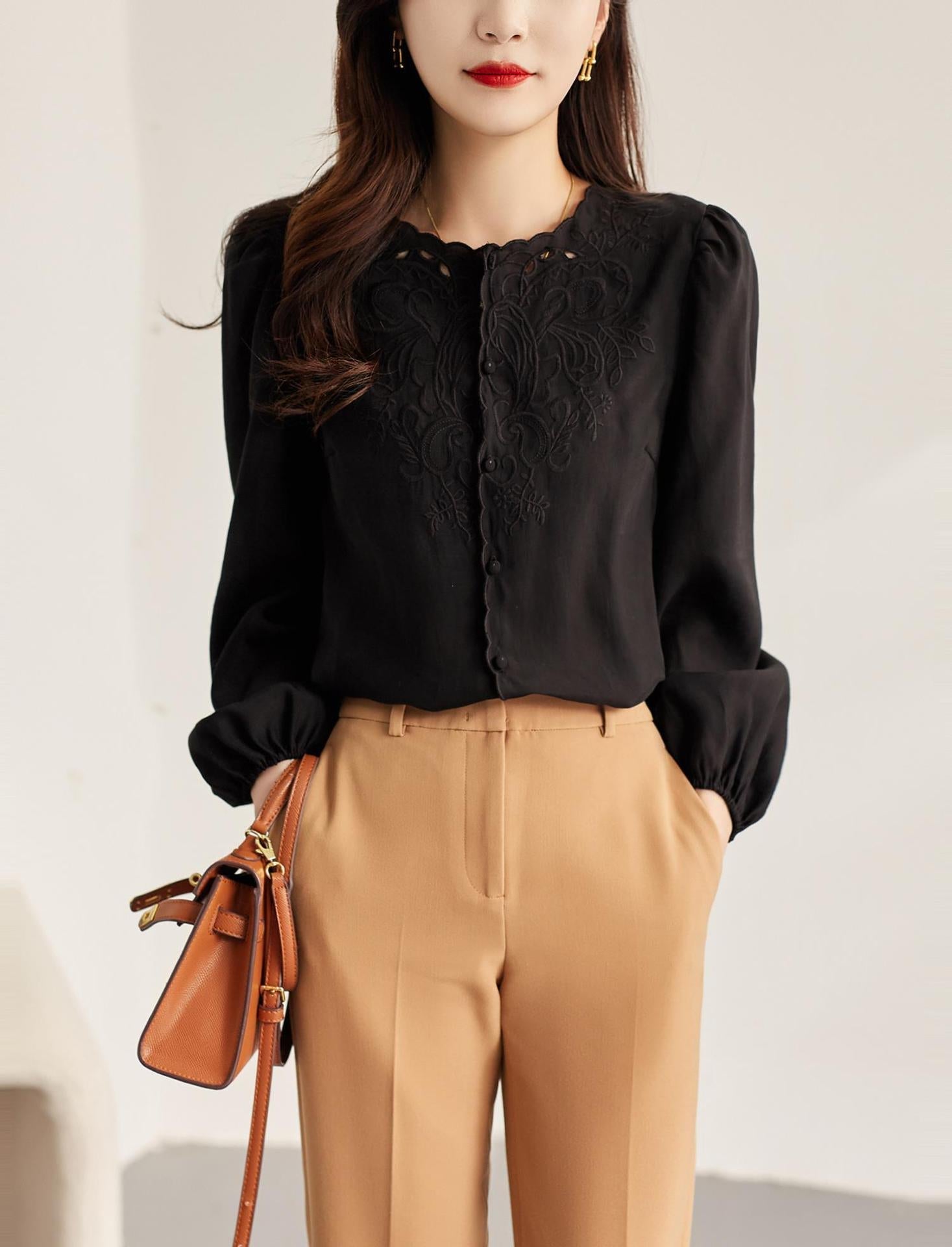 A woman wearing a black long-sleeve blouse with delicate floral embroidery on the chest and puffed sleeves, paired with tan trousers and holding a brown handbag.