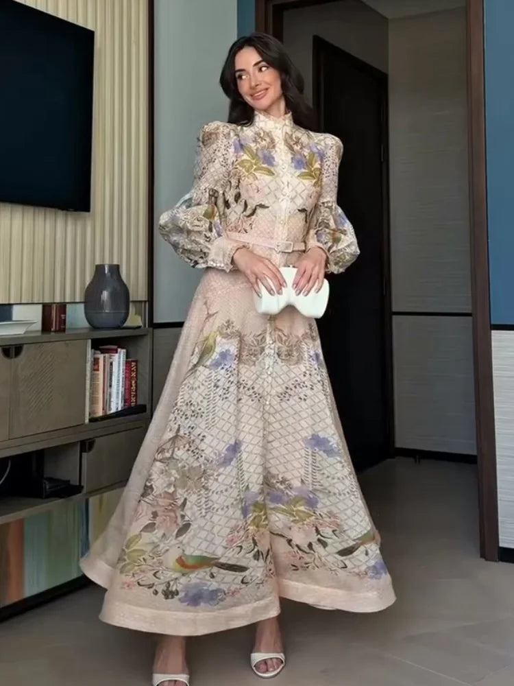 A woman models a long-sleeved, high-neck maxi dress with a beige base and intricate multicolor floral and bird embroidery, holding a white clutch in an indoor setting.