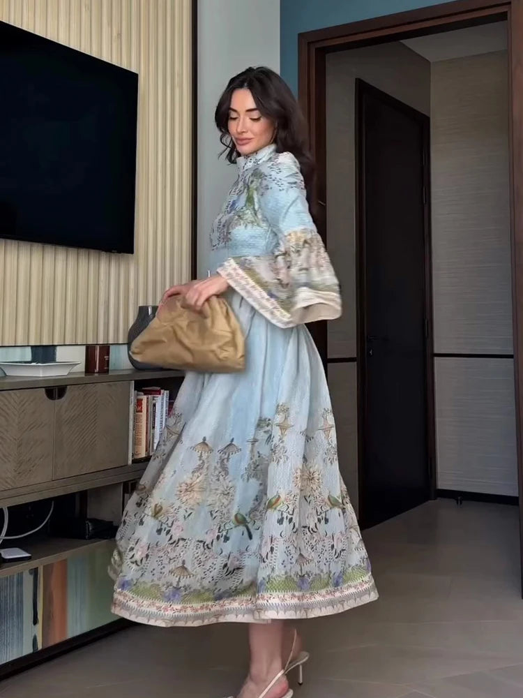 A woman models a light blue traditional dress with intricate floral embroidery, holding a tan leather clutch bag in an indoor setting.