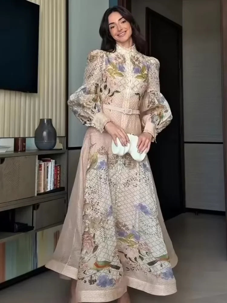 A woman wearing a long beige embroidered dress with floral and bird motifs, holding a white clutch in an indoor setting.