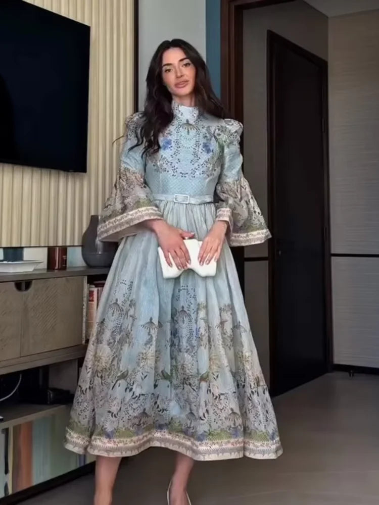 A woman stands indoors wearing a light blue midi dress with ornate floral embroidery, puffed sleeves, and a gathered waist. She is holding a small white clutch in a modern room with a TV, cabinet, and doorway visible.