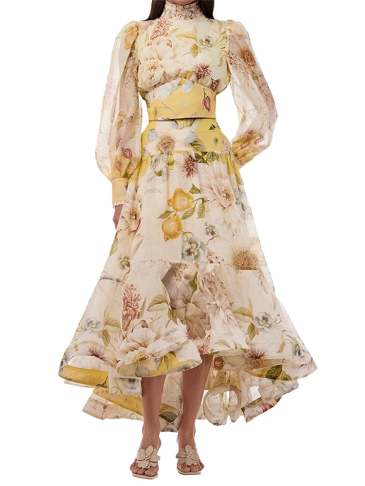 A model wearing a high-low midi dress with a yellow and beige floral print, featuring long sheer puff sleeves and a matching waist belt, set against a plain white background.