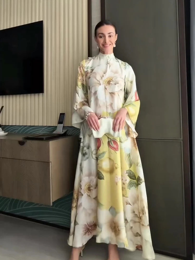 A woman wearing a long-sleeved, high-neck maxi dress with a large floral print in cream, yellow, and green, standing in a modern indoor setting.