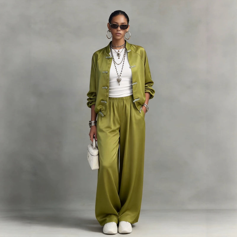 A model stands against a textured gray studio backdrop wearing a matching olive green satin jacket and wide-leg trousers set with a white ribbed top and white sneakers.