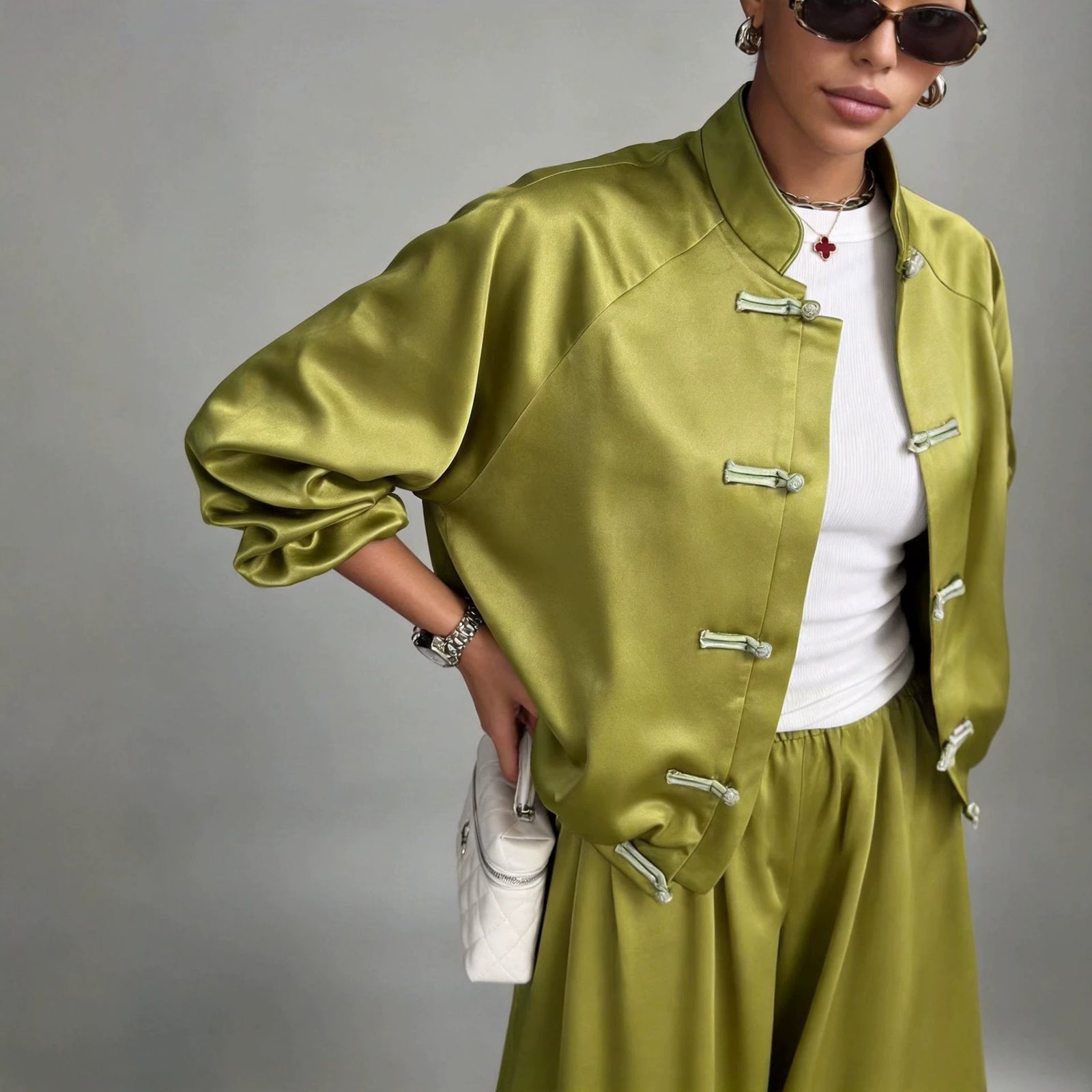 A model wearing a shiny olive green satin jacket and matching pants with silver-tone frog closures, styled over a white top and sunglasses, against a plain gray studio background.