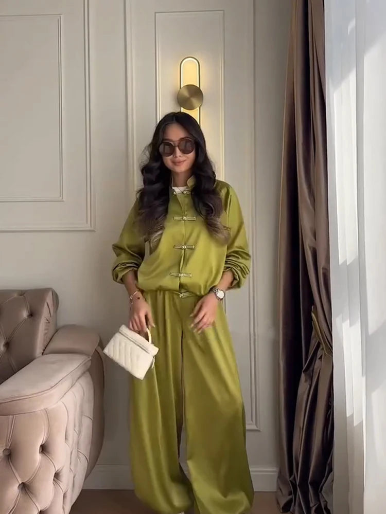 A woman wearing a shiny olive green two-piece jumpsuit with traditional Chinese frog closures, standing in a decorated room with a tufted sofa and curtains.