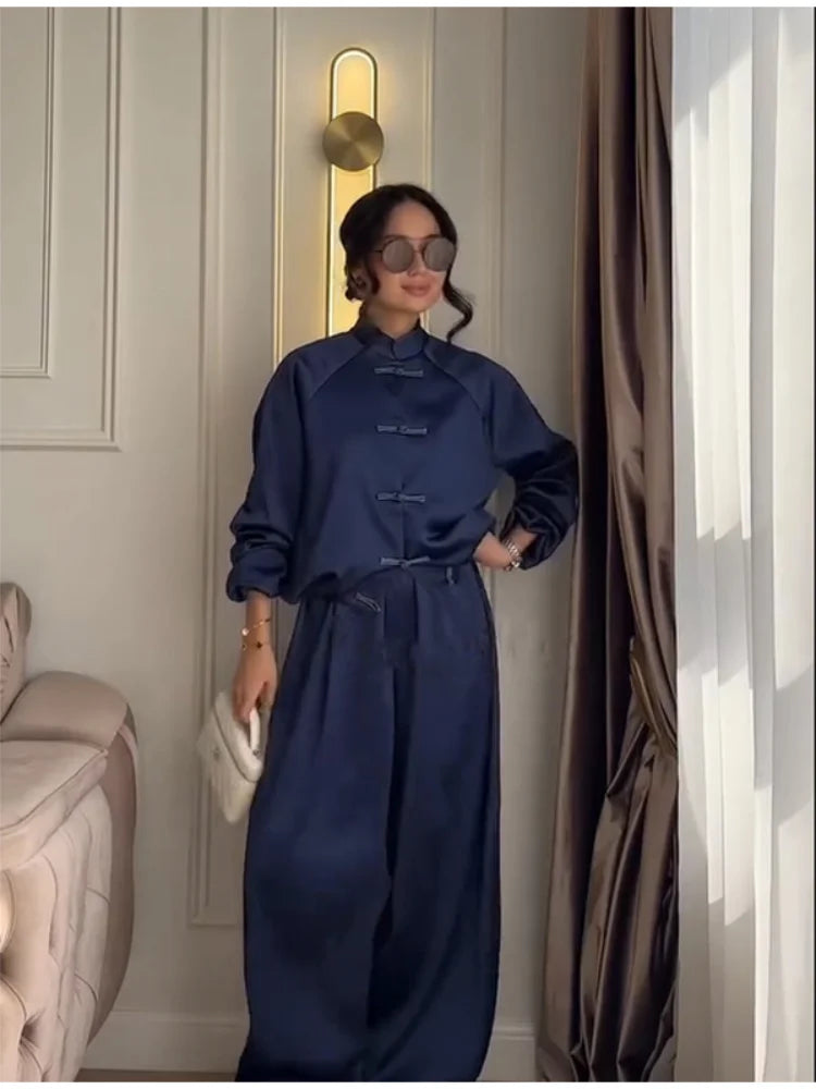 A woman wearing a navy blue satin two-piece outfit with a high collar and traditional frog closures, standing in an indoor setting with curtains and a wall lamp visible.