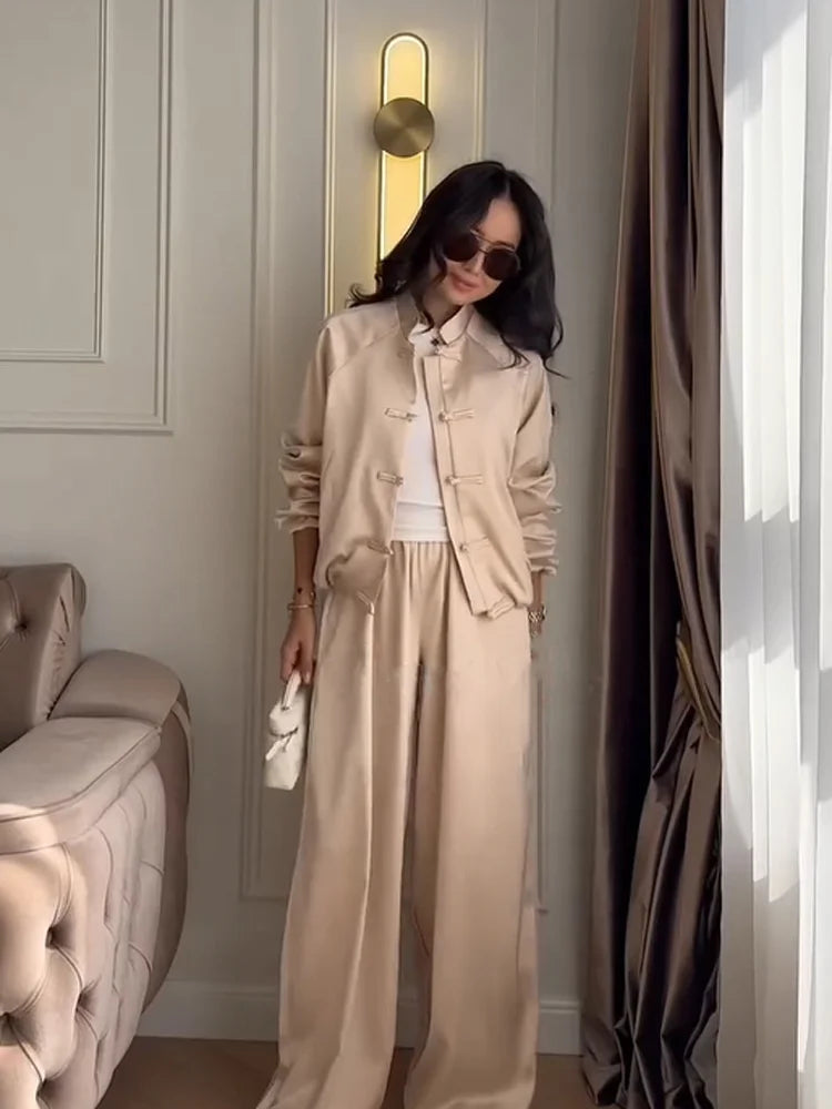 A woman models a beige satin-like two-piece suit consisting of a cropped jacket with traditional Chinese frog closures and matching wide-leg trousers, styled with a white top and sunglasses in an elegant room.