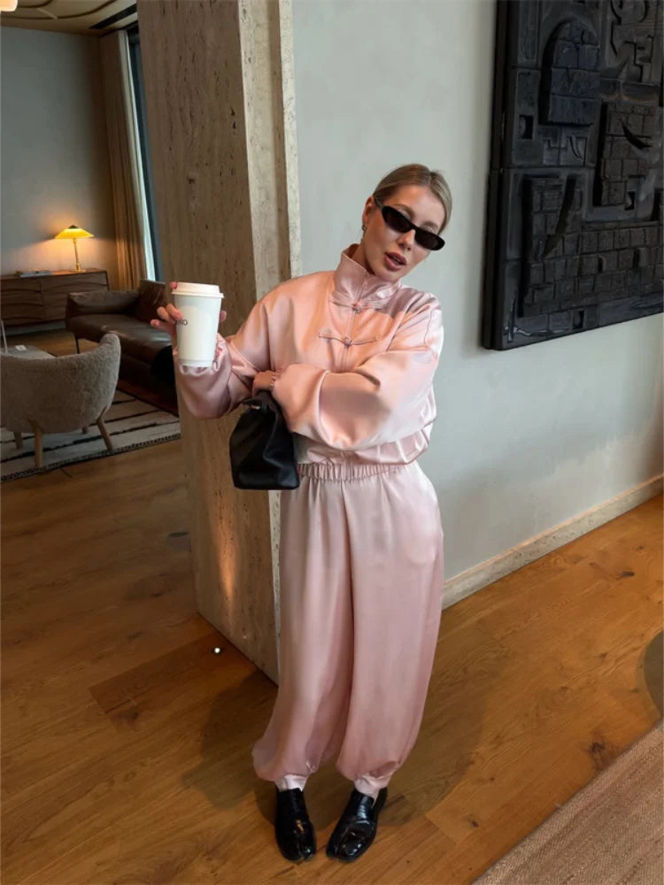 A woman wearing a light pink matching lounge set with a high-neck long-sleeve top and wide-leg pants, styled with black sunglasses, a black handbag, and black loafers, standing in a modern indoor setting with wood floors and walls.