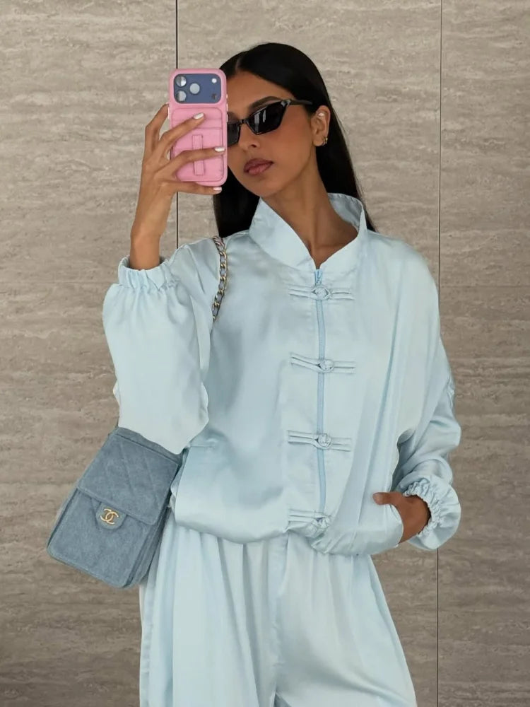 A woman takes a mirror selfie wearing a light blue traditional Chinese-style jacket and matching pants, holding a pink phone case, and carrying a blue quilted shoulder bag against a beige tiled wall.