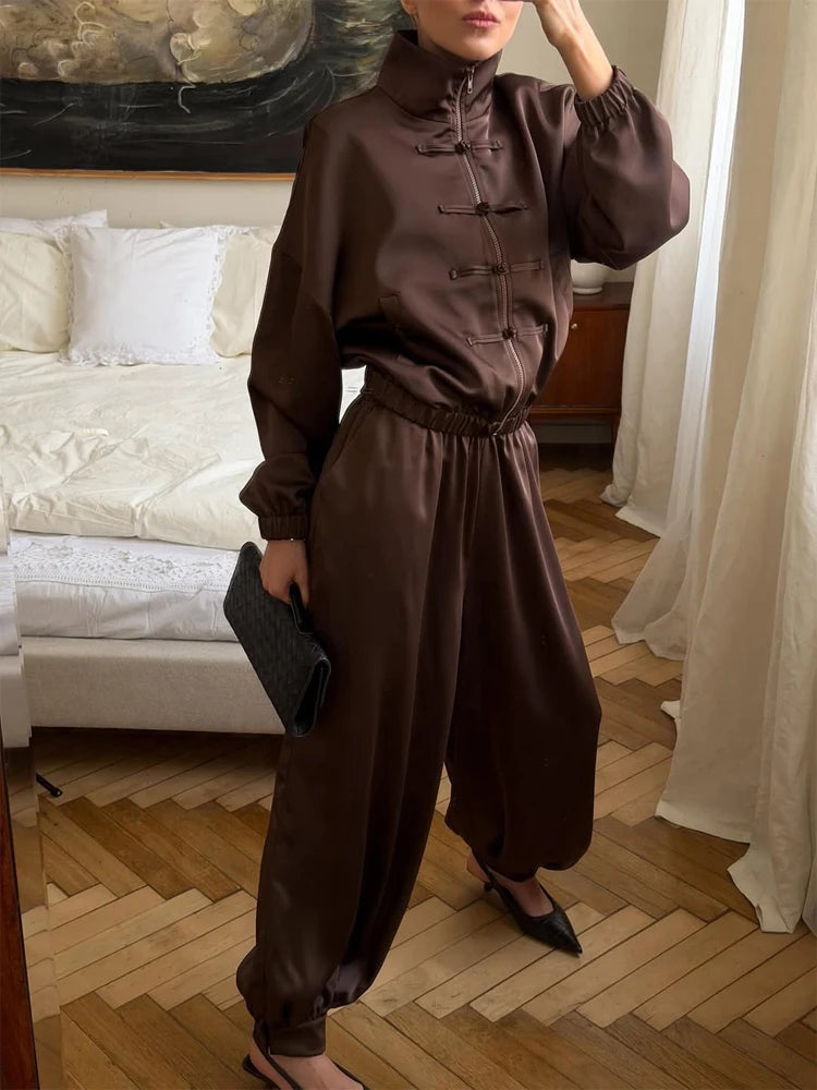 A woman models a dark brown satin two-piece set featuring a high-neck, long-sleeve top with traditional Chinese frog closures and matching wide-leg harem pants, styled in a bedroom setting.
