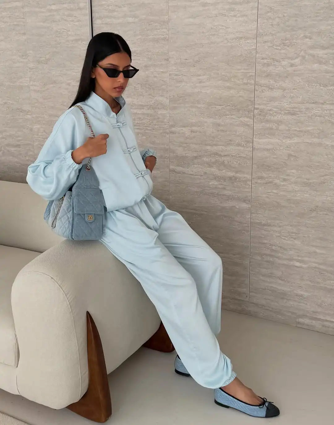 A woman wearing a light blue two-piece tracksuit with traditional Chinese-style frog closures, sitting on a cream-colored sofa. She is wearing black sunglasses, a light blue quilted shoulder bag, and light blue and black flat shoes against a beige textured wall.