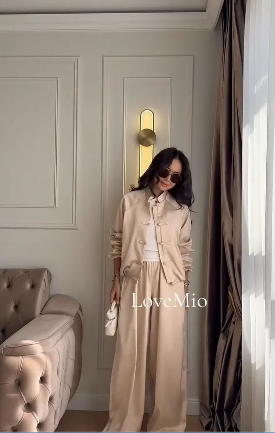 A woman wearing a beige two-piece lounge set consisting of a button-up shirt and matching wide-leg pants, standing in a modern room with a tufted sofa, wall sconce, and curtains.