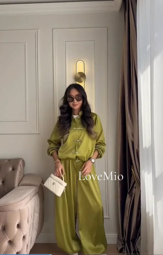 A woman wearing a matching olive green two-piece outfit with wide-leg pants and long sleeves, standing in a decorated room with a tufted sofa, curtains, and a wall light. She is holding a small white handbag, and the text 'LoveMio' is overlaid on the image.