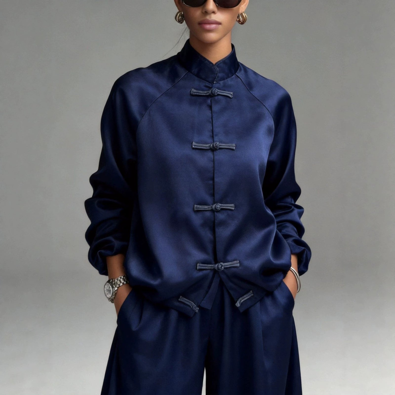 A model wearing a navy blue satin long-sleeve top with traditional Chinese frog closures, paired with matching pants, set against a neutral gray studio background.