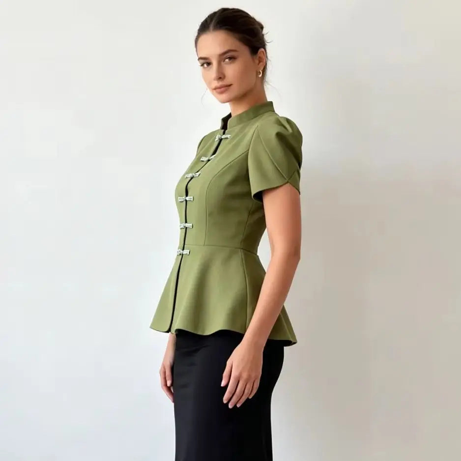 A woman modeling a sage green short-sleeve peplum top with black trim and white decorative buttons, paired with a black skirt, against a plain light gray background.