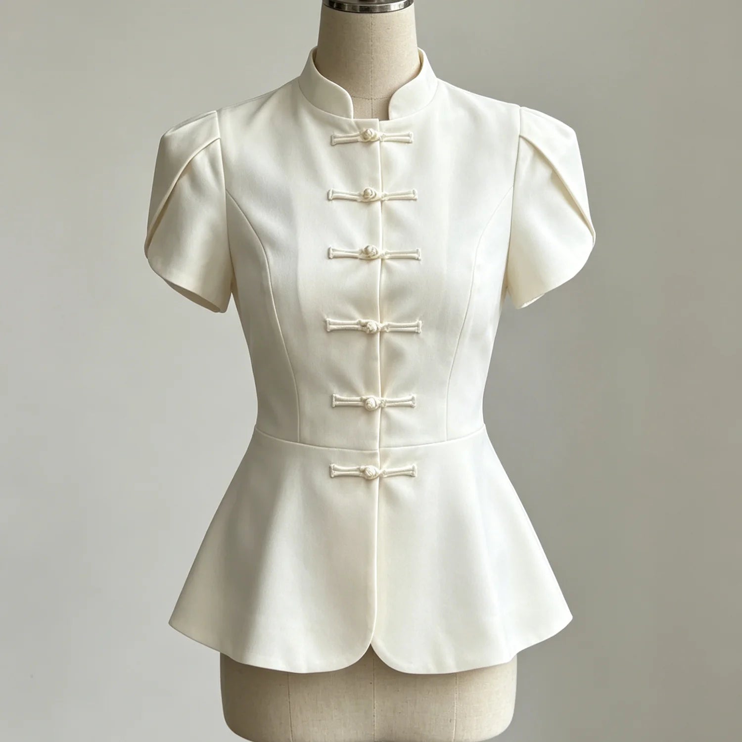 An ivory short-sleeve peplum top with traditional Chinese frog closures, displayed on a cream-colored dress form mannequin against a neutral light gray background.