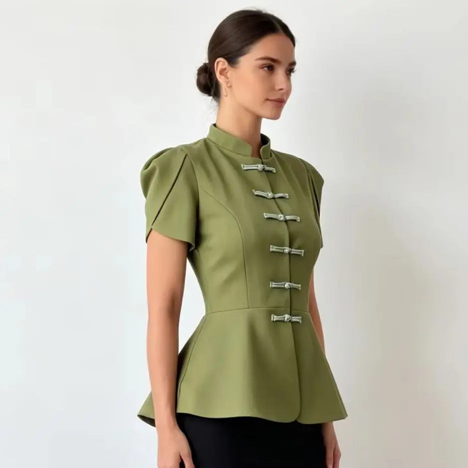 A woman wearing a sage green short-sleeve blouse with silver-toned decorative frog closures down the front, paired with black bottoms, photographed against a plain light background.