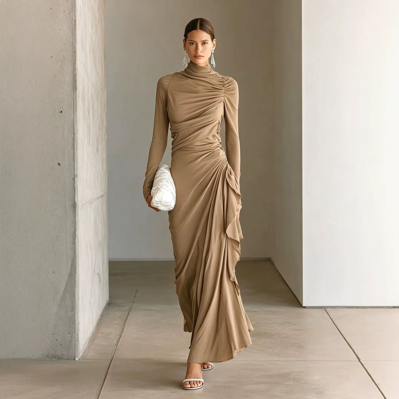 A woman models a draped, long-sleeve maxi dress in a taupe-brown color, styled with a white textured clutch and white heeled sandals in a minimalist concrete-walled corridor.