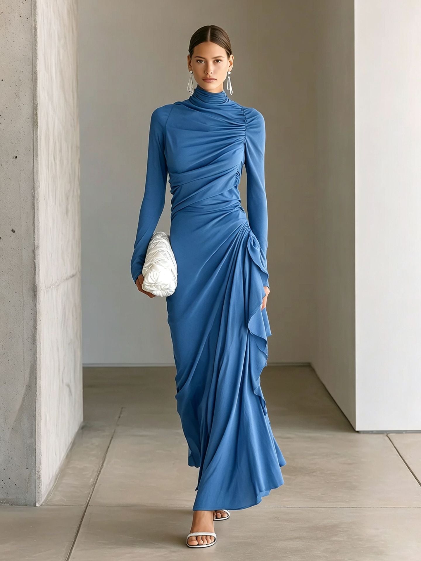 A woman models a long-sleeved, high-neck blue maxi dress with dramatic draped and gathered details. She is holding a white textured clutch and wearing silver sandals, standing in a minimalist indoor setting with concrete pillars and neutral walls.