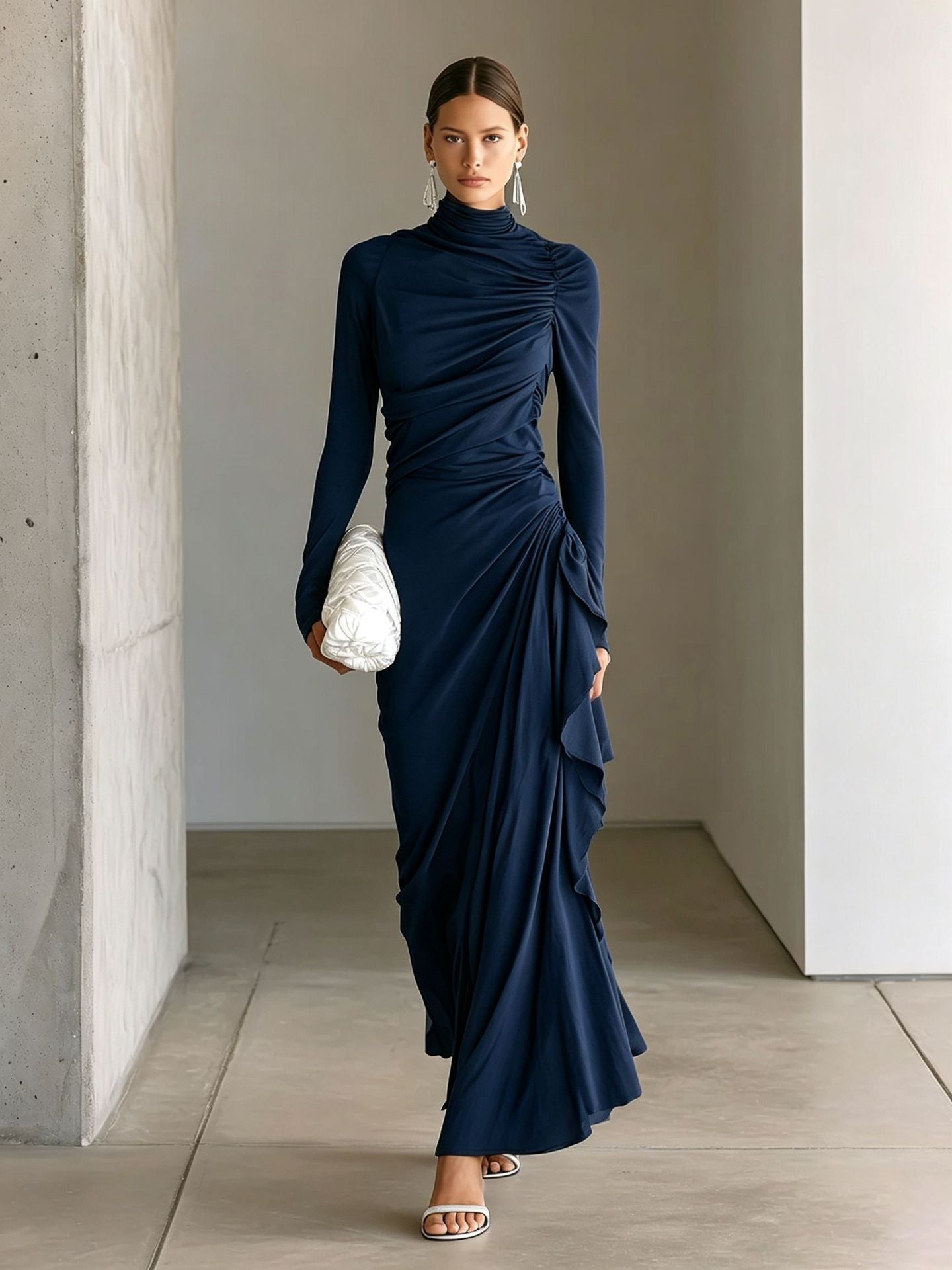 A woman models a navy blue long-sleeve maxi dress with dramatic ruching and draped details, styled with a white textured clutch and silver sandals in a minimalist concrete corridor.