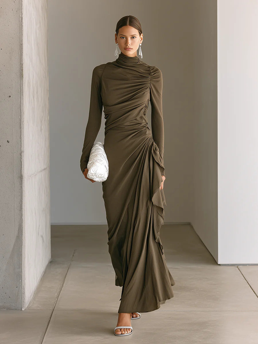 A woman models a long-sleeved, high-neck, ruched maxi dress in a dark olive brown color. She is holding a white textured clutch and wearing silver sandals, standing in a minimalist corridor with concrete walls.