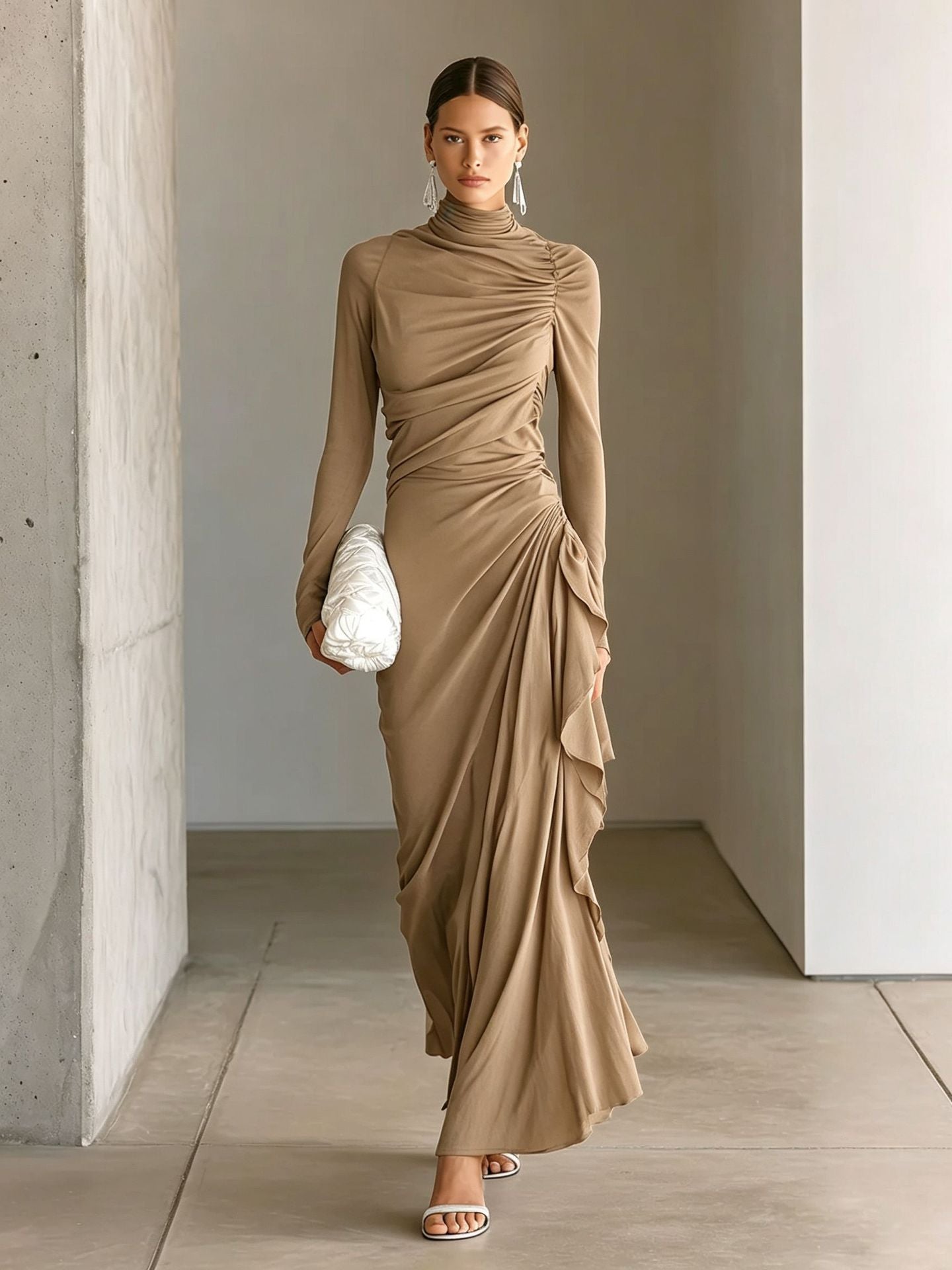 A woman models a long-sleeved, high-neck, ruched maxi dress in a warm taupe-brown shade. She is walking in a minimalist corridor with concrete walls and light-colored flooring, holding a white textured clutch.