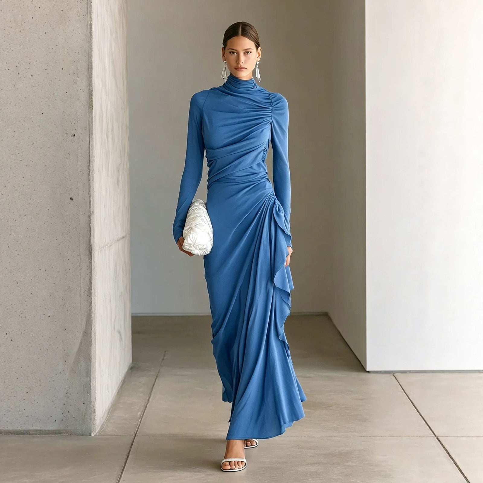 A woman models a long-sleeve, high-neck, blue ruched maxi dress in a minimalist indoor setting with concrete and plaster walls.