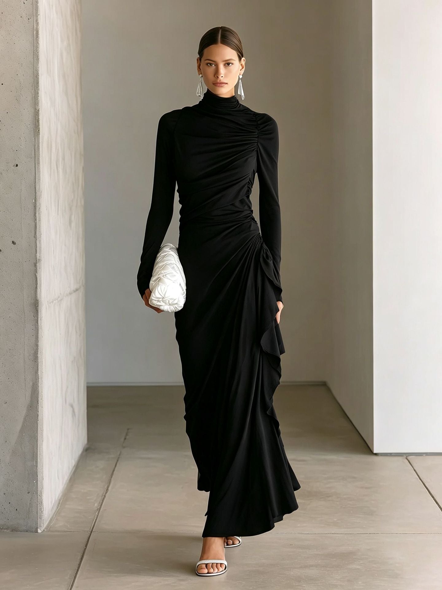 A model walks down a minimalist concrete corridor wearing a long-sleeved black draped dress with a high ruffled collar, holding a white textured clutch.