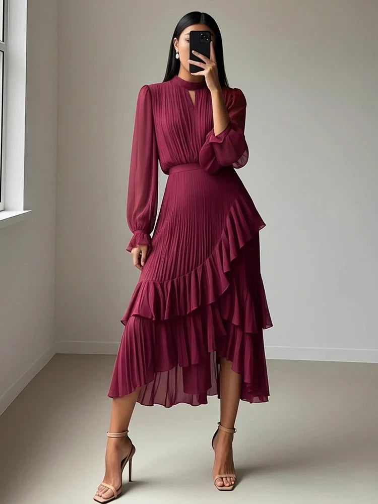 A woman modeling a long-sleeved, high-neck midi dress in deep burgundy with a pleated ruffled skirt, taking a mirror selfie in a minimalist room.