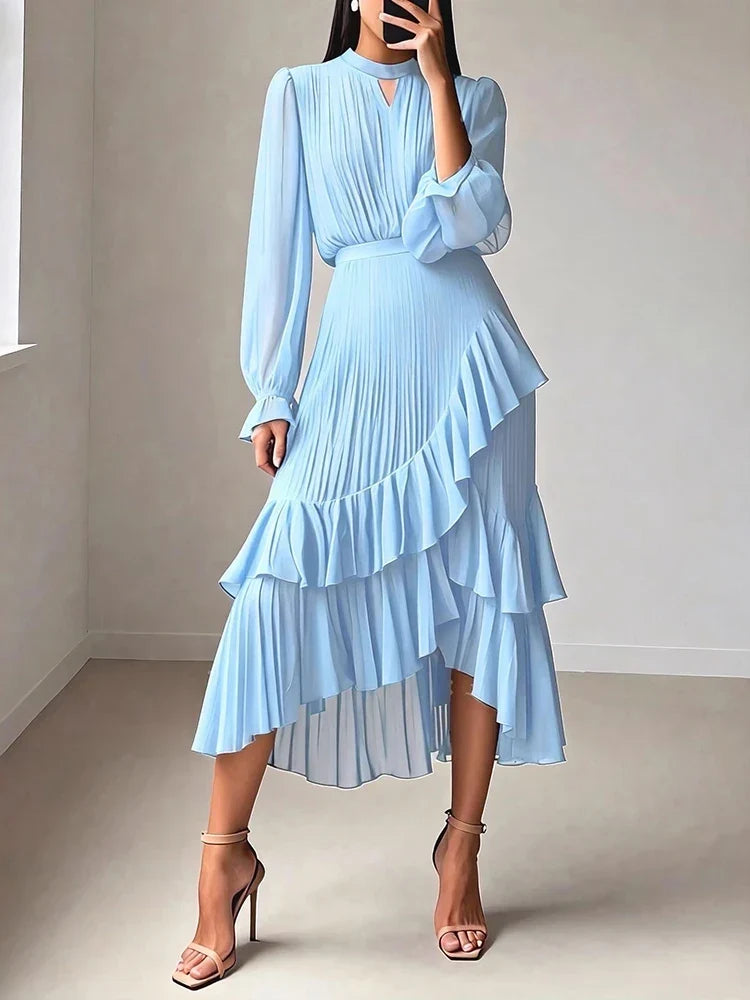 A woman wearing a light blue midi dress with long sheer puff sleeves, a pleated high-waisted ruffled skirt, and a keyhole cutout at the neckline, paired with nude high-heeled sandals in a minimalist indoor setting.