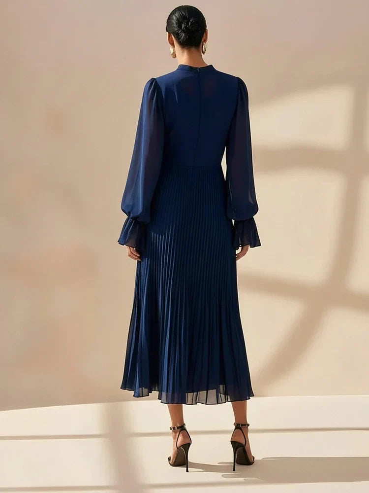 A woman viewed from behind wearing a navy blue midi dress with long sheer puff sleeves and a pleated skirt, paired with dark strappy heels, against a warm beige wall with soft window shadows.