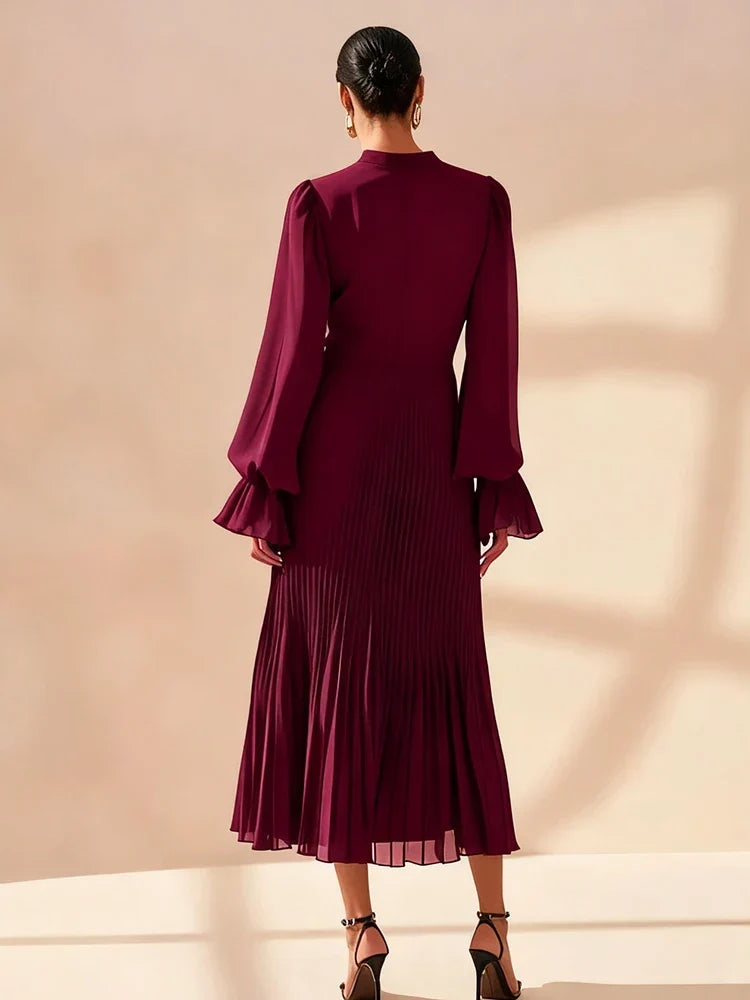 A woman shown from the back wearing a deep burgundy midi dress with long ruffled sleeves and a pleated skirt, standing against a warm beige studio background.