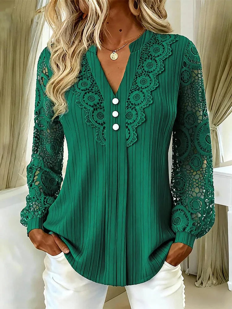 A woman wearing a green long-sleeve blouse with lace panels on the sleeves and neckline, featuring a V-neckline with three white buttons, paired with white pants.