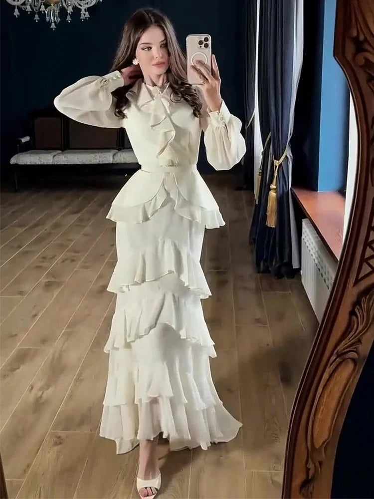 A woman takes a mirror selfie wearing a long cream-colored ruffled dress with a high neck, sheer long sleeves, and a tiered skirt, set in a room with wooden floors and dark blue curtains.