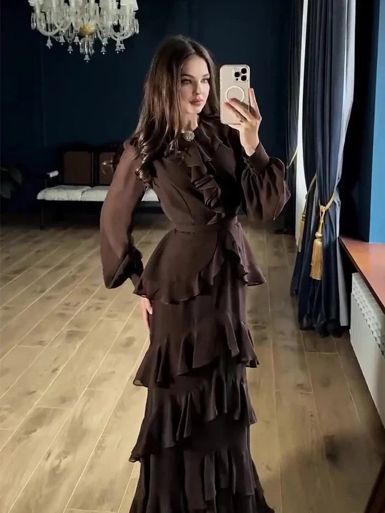 Woman taking a mirror selfie wearing a dark brown ruffled dress in a room with wooden flooring and blue walls.
