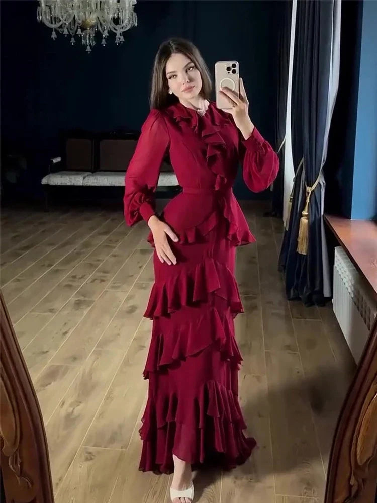 A woman takes a mirror selfie wearing a long-sleeved, deep burgundy ruffled maxi dress in a room with a wooden floor, dark blue walls, and curtains.