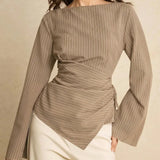 A woman wearing a taupe pinstriped long-sleeve top with a wrap-style waist and cream-colored pants, photographed against a warm beige studio background.