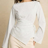 A woman wearing a white long-sleeve wrap-style blouse with thin vertical navy pinstripes and cream pants, photographed against a warm beige studio background.