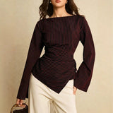 A woman wearing a dark burgundy long-sleeve top with thin vertical red and gold pinstripes, paired with cream-colored pants, holding a small brown handbag against a neutral beige background.