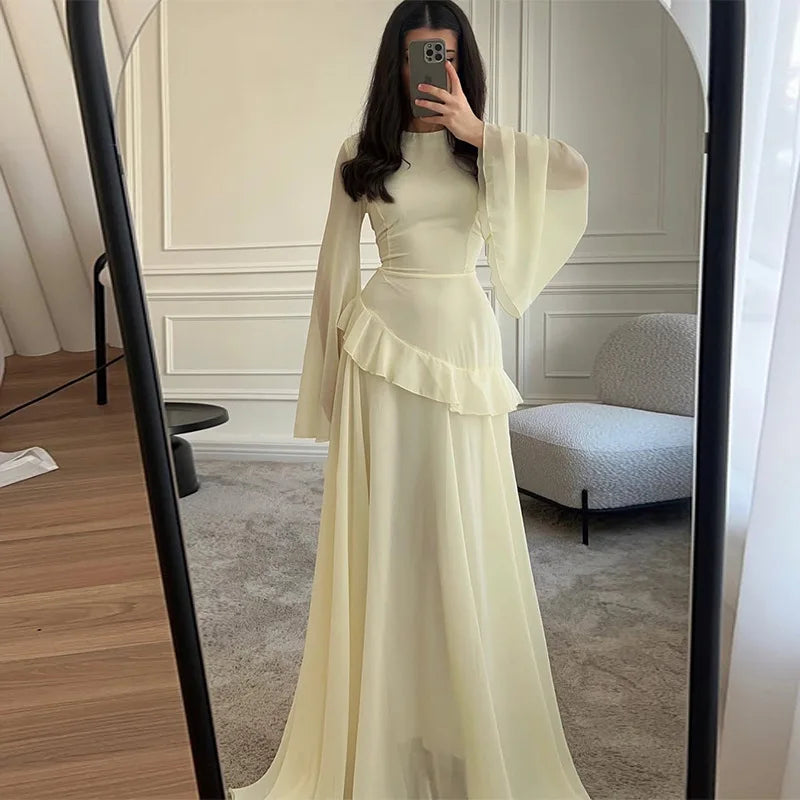 Woman wearing a long, light-colored dress with sheer sleeves in a room.