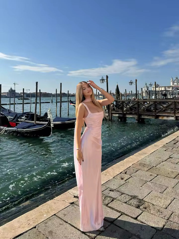 A woman posing on a stone pier by a canal with gondolas and water in the background, wearing a light pink sleeveless maxi dress.