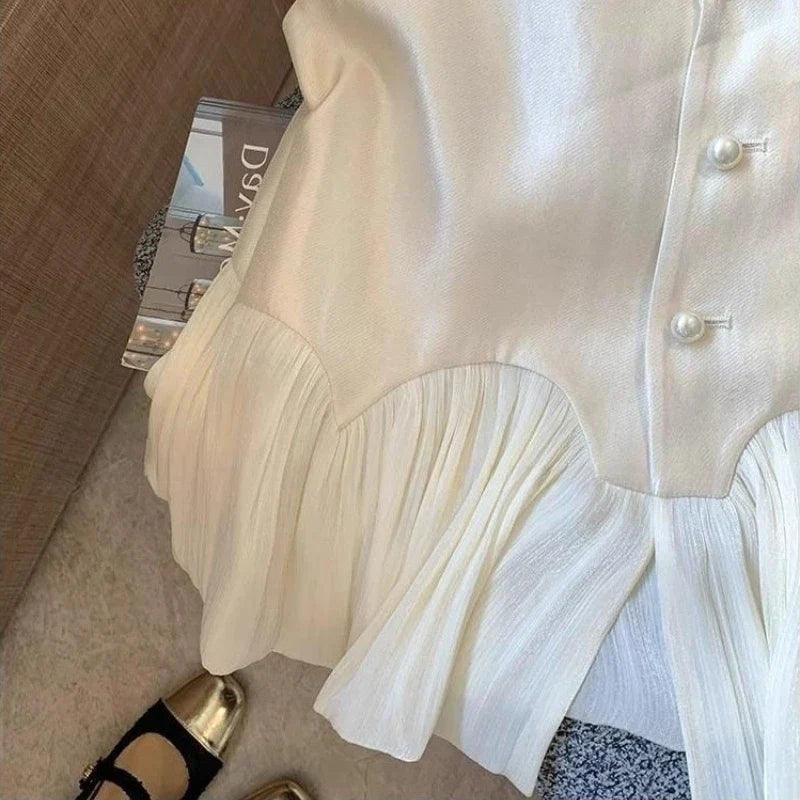 An off-white or cream-colored blouse with a pleated ruffled hem and pearl-like buttons, shown in a styled flat-lay setting next to a book and shoes.