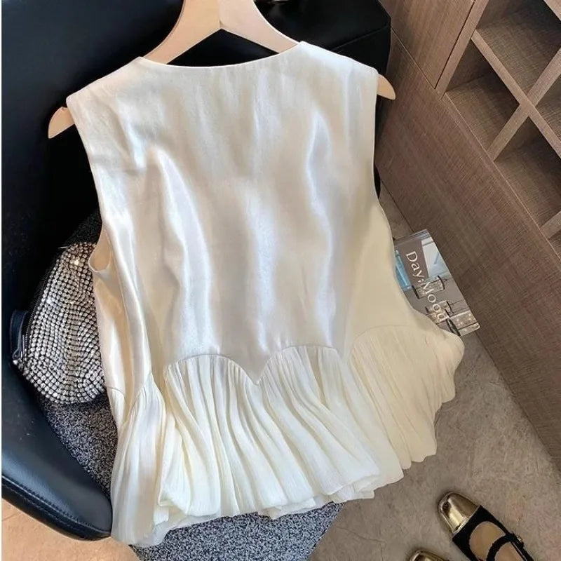 A sleeveless white top with a pleated ruffled hem, hanging on a wooden hanger on a black chair next to a silver patterned clutch.