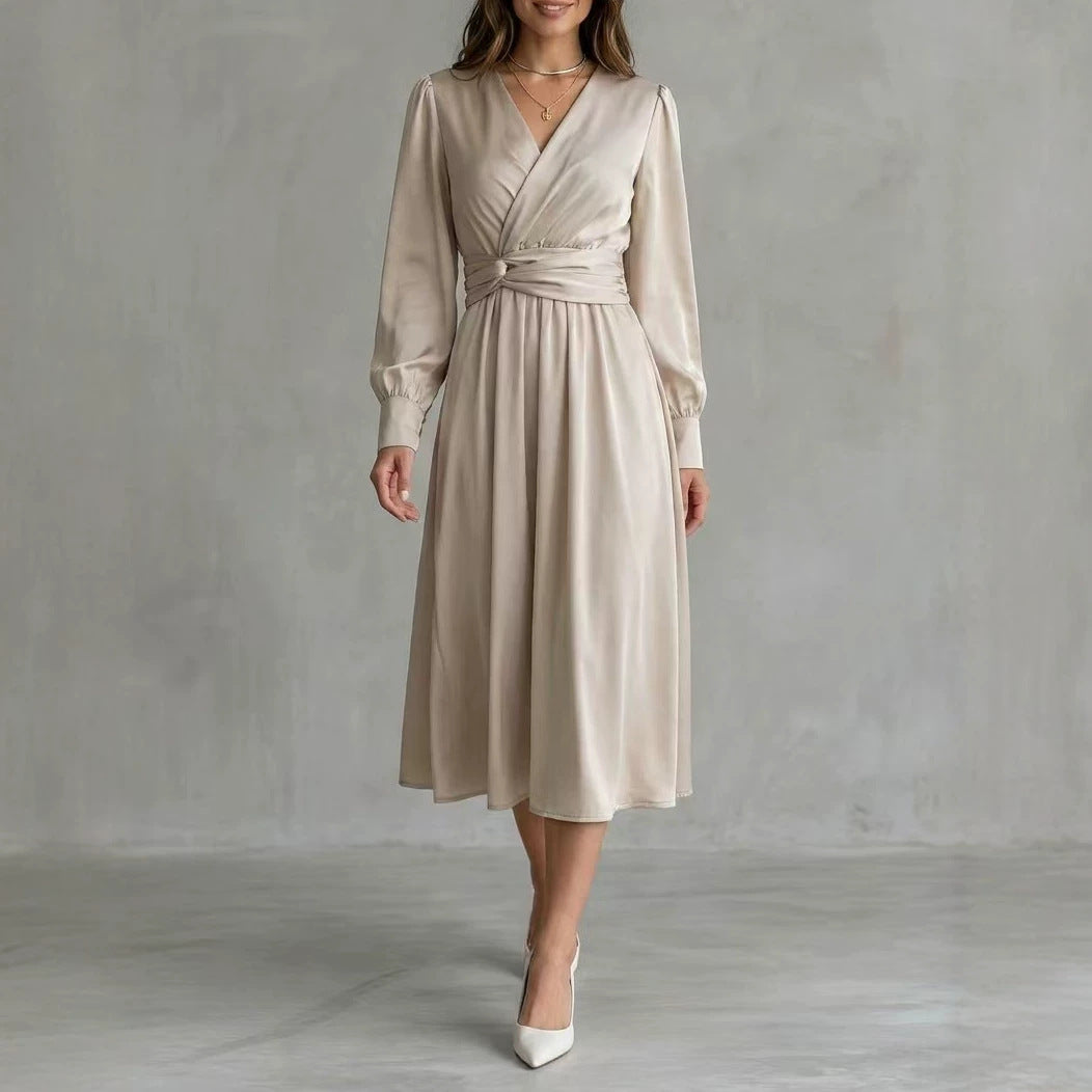 A woman models a light beige satin midi dress with long sleeves, a V-neckline, and a matching fabric tie belt at the waist, paired with white pointed-toe heels against a textured gray studio backdrop.